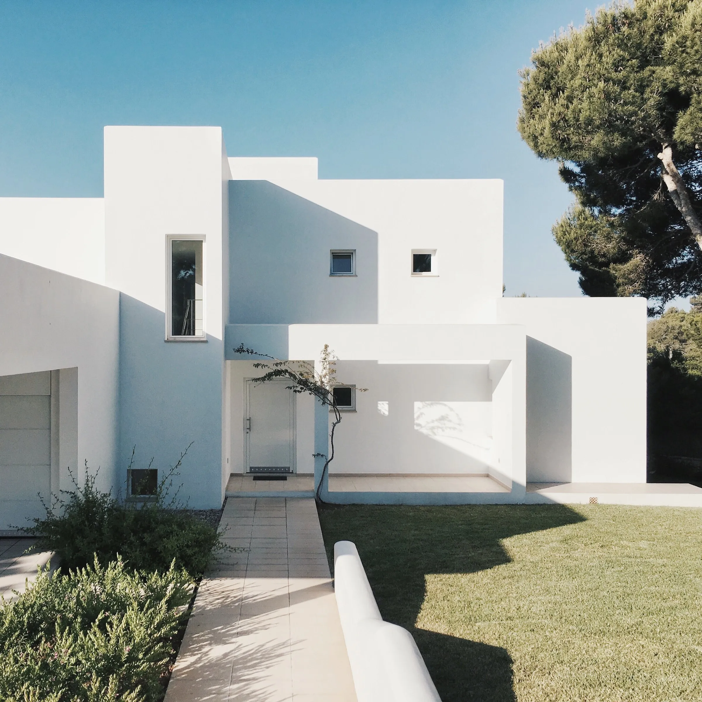 White cubic house with narrow windows beside a tall pine tree under a blue sky