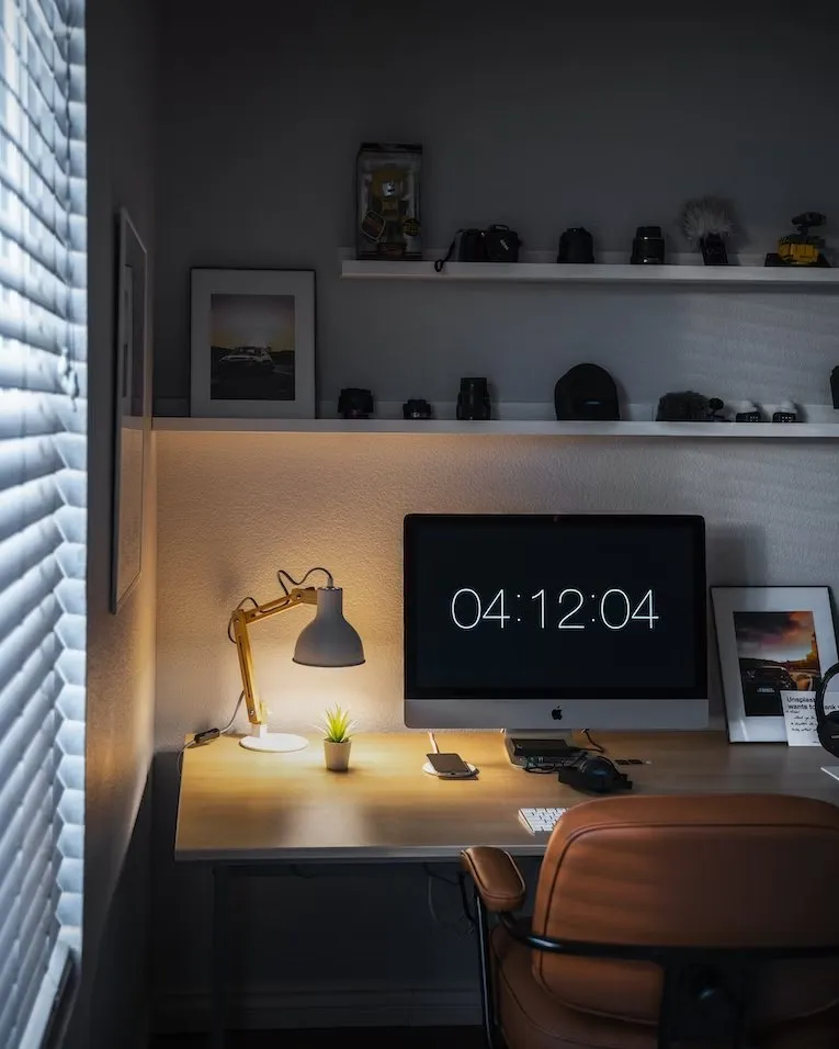 Dark studio desk with a desk lamp, a computer showing the time and shelves of cameras and models